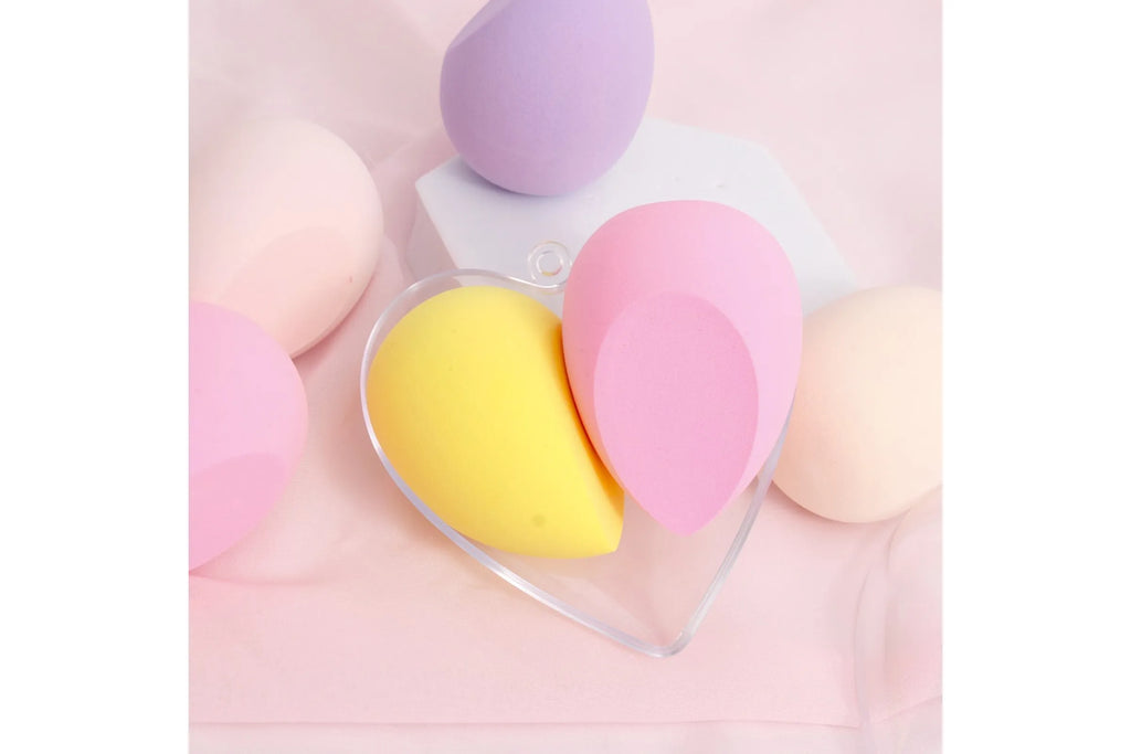 Heart-shaped clear plastic case with yellow and pink makeup beauty blenders surrounded by assorted pastel sponges