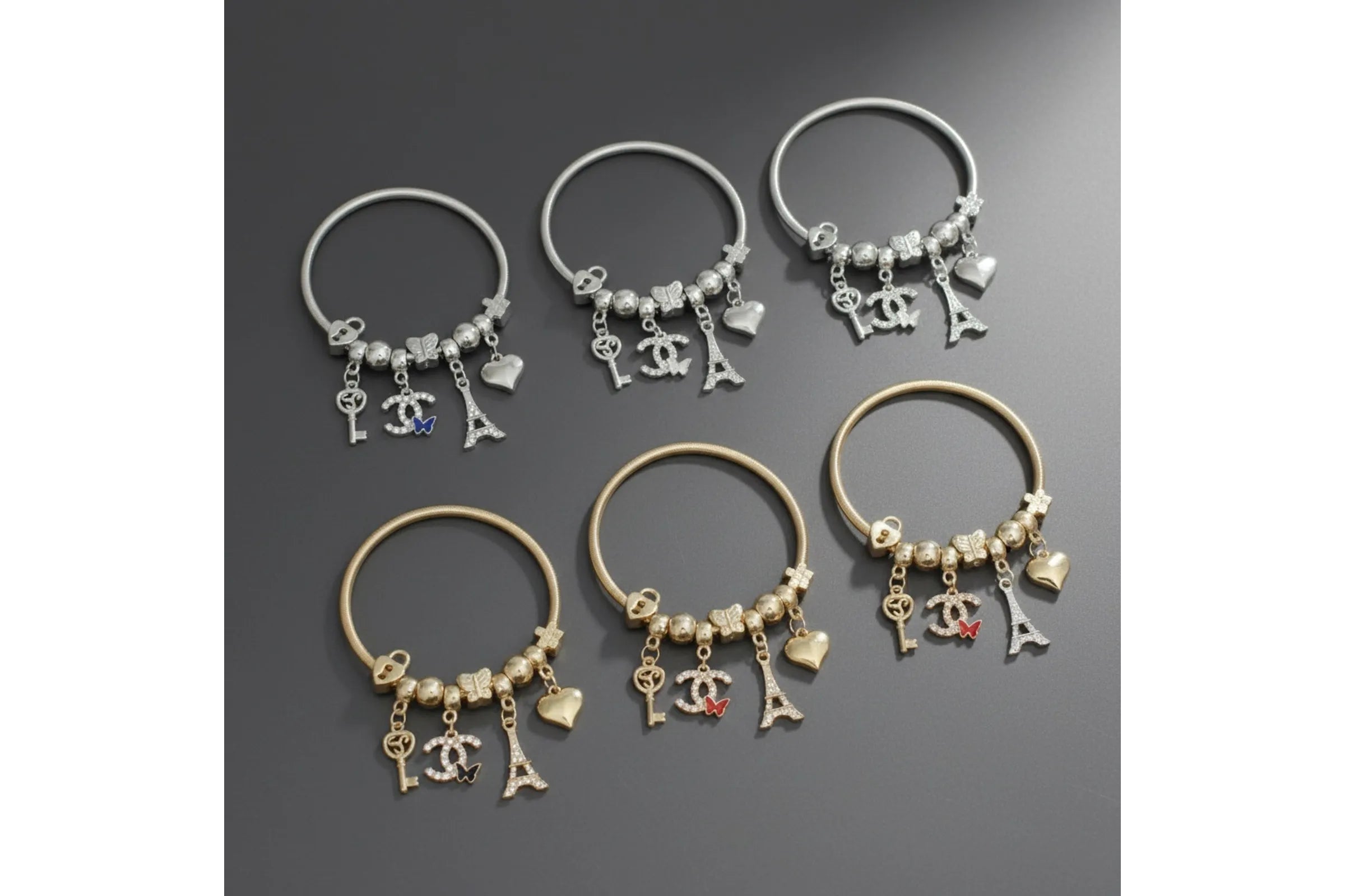 Six metal jump coil stretch bracelets arranged in two rows of three on a dark gray surface. The top row shows three silver tone bracelets and the bottom row shows three gold tone bracelets, each featuring assorted charms including a key, interlocking letter, Eiffel Tower, puffy heart, and enamel butterfly in varying colors.