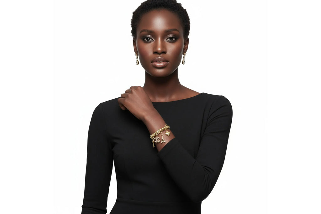 A woman in a black long-sleeve boat-neck dress poses against a white background with her right hand raised to her shoulder. She wears a gold tone metal jump coil stretch bracelet featuring interlocking letter, Eiffel Tower, puffy heart, and key charms, and drop crystal earrings.