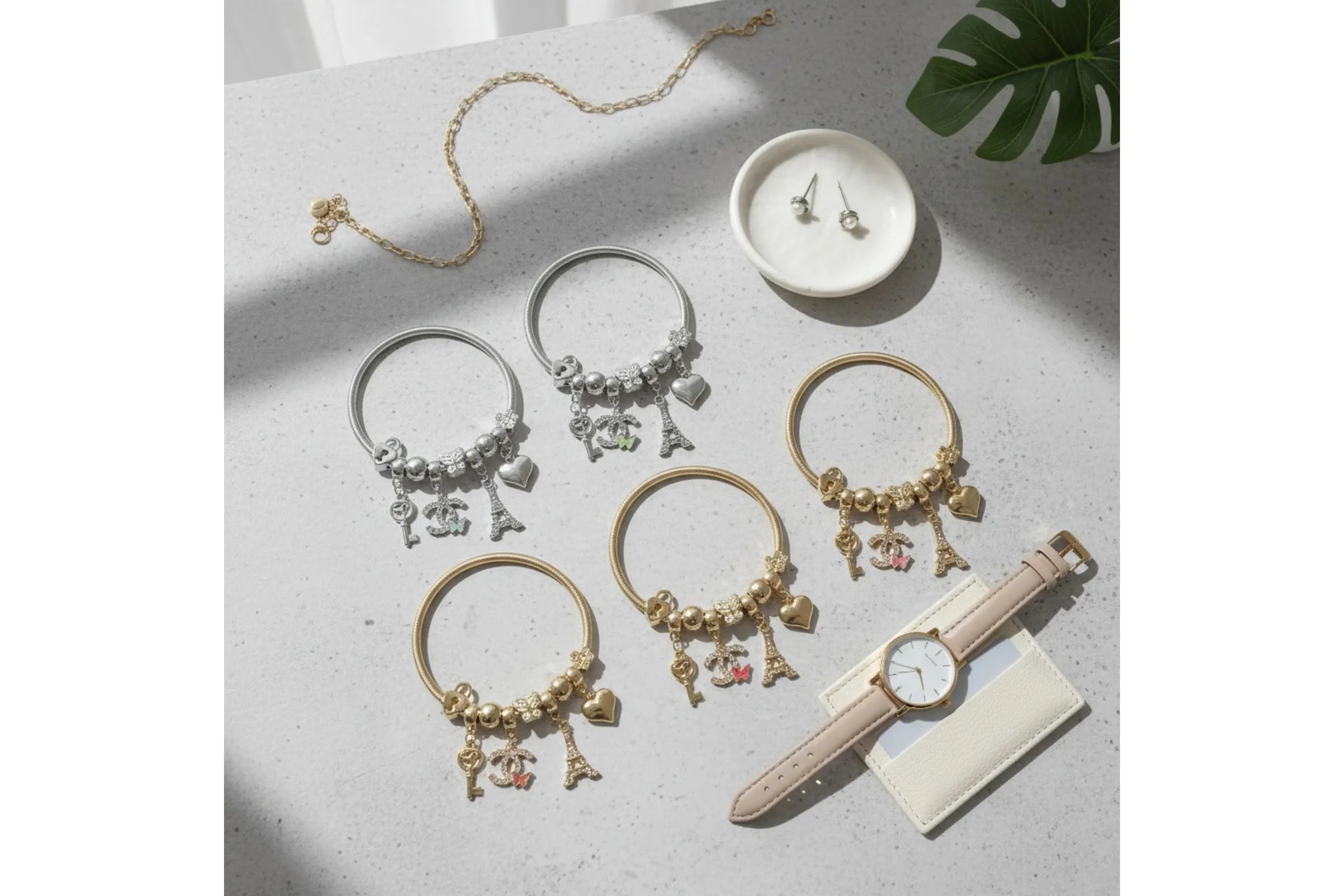 A styled flat-lay of jewelry and accessories on a light gray surface with natural shadows. The set includes multiple beaded bracelets in silver and gold with Eiffel Tower and heart charms, a delicate gold chain necklace, a pair of silver stud earrings in a small white dish, a beige leather watch strap, and a cream card holder.