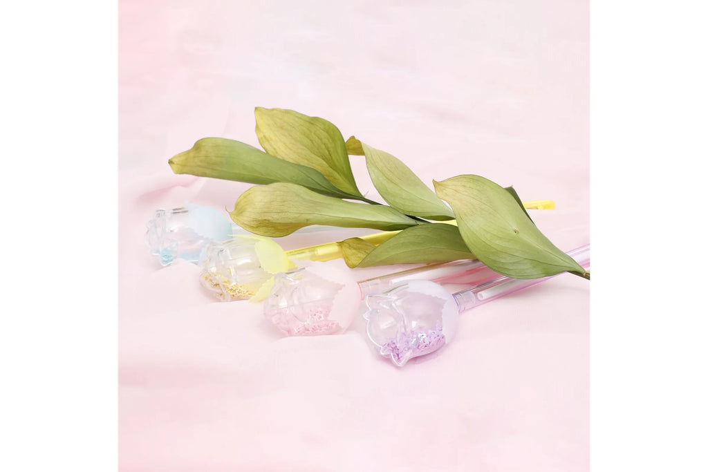 Three pastel plastic rose bud pens with clear confetti-filled rose bud toppers in blue, yellow, and lavender arranged alongside a green leafy stem on a soft blush pink fabric surface, styled to resemble a bouquet of flowers.