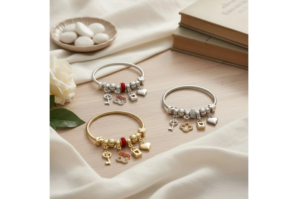 Overhead flat lay on a blush pink wood surface with white silk fabric. Three metal jump coil stretch bracelets arranged in a loose cluster — one silver tone with red rhinestone rondelle bead, one silver tone with clear white rhinestone rondelle bead, and one gold tone with red rhinestone rondelle bead — each featuring dangling key, quatrefoil clover, heart lock, and puffy heart charms. Styled with a white rose with green leaves, a small ceramic dish with white oval stones, and two stacked tan hardcover book