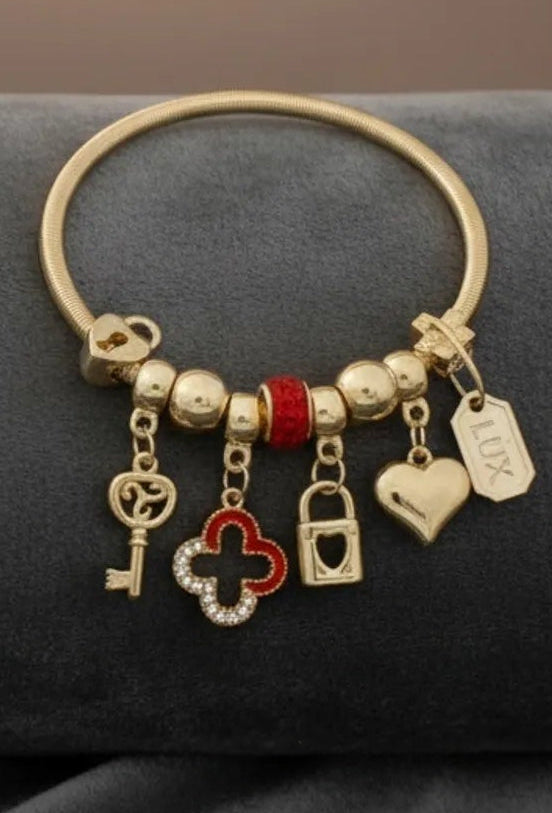 Close-up of a gold tone metal jump coil stretch bracelet displayed on a dark gray velvet surface featuring a red rhinestone rondelle slide bead, gold ball beads, and dangling key, red and clear rhinestone quatrefoil clover, heart lock padlock, puffy heart, and small rectangular tag charm in gold tone.