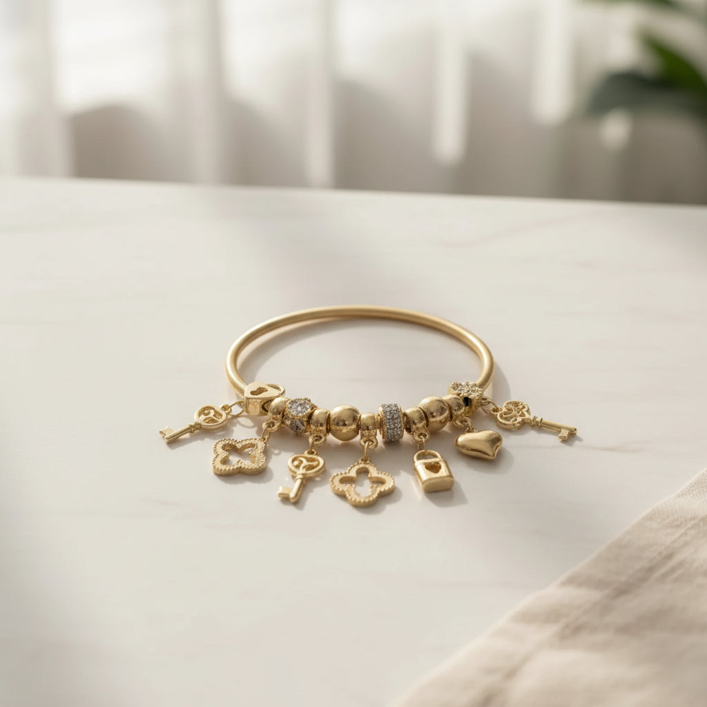 Gold tone metal jump coil stretch bracelet laid flat on a white marble surface near a sheer curtained window, featuring clear white rhinestone rondelle spacer beads, gold ball beads, and dangling key, open quatrefoil clover, heart lock padlock, and puffy heart charms in gold tone. A green plant is softly visible in the upper right background.
