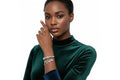 A woman with dark skin wearing an emerald green velvet turtleneck poses against a white background with her right hand raised to her cheek. She wears a silver tone metal jump coil stretch bracelet with a red rhinestone rondelle slide bead and dangling key, rhinestone quatrefoil clover, heart lock, and puffy heart charms on her left wrist.
