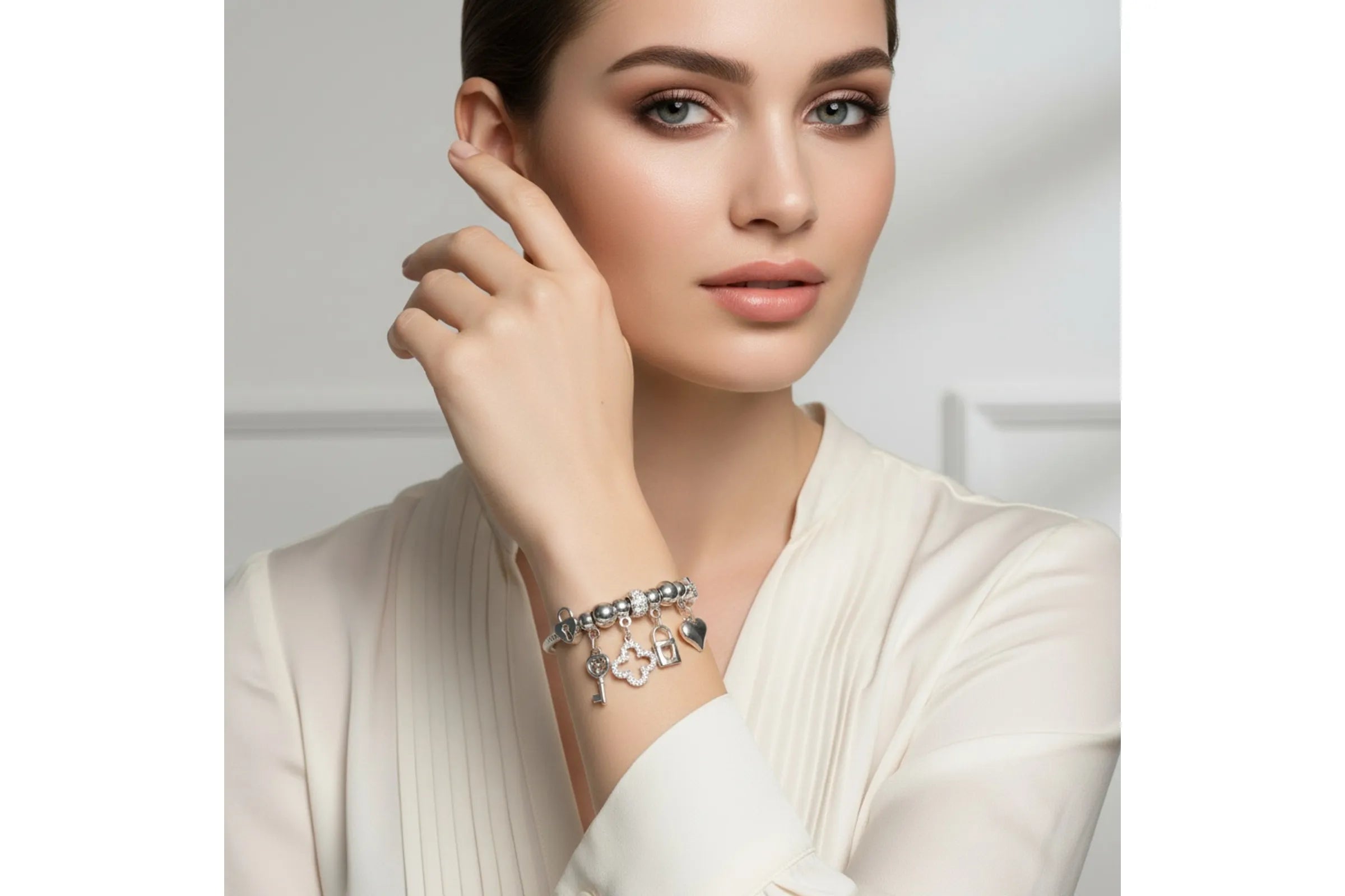 A woman with light skin and dark hair pulled back poses against a soft white background with her right hand raised to her cheek. She wears a silver tone metal jump coil stretch bracelet with a red rhinestone rondelle slide bead and dangling key, rhinestone quatrefoil clover, heart lock, and puffy heart charms, styled with a cream structured blazer.