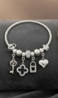 Close-up of a silver tone metal jump coil stretch bracelet displayed on a dark gray surface featuring clear white rhinestone rondelle spacer beads, silver ball beads, and dangling key, quatrefoil clover, heart lock, and puffy heart charms in silver tone.