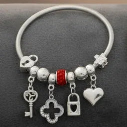 Close-up of a silver tone metal jump coil stretch bracelet laid flat on a dark gray surface featuring a red rhinestone rondelle slide bead, silver ball spacer beads, and dangling key, open quatrefoil clover, heart lock padlock, and puffy heart charms in silver tone.