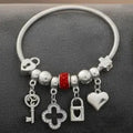 Close-up of a silver tone metal jump coil stretch bracelet laid flat on a dark gray surface featuring a red rhinestone rondelle slide bead, silver ball spacer beads, and dangling key, open quatrefoil clover, heart lock padlock, and puffy heart charms in silver tone.