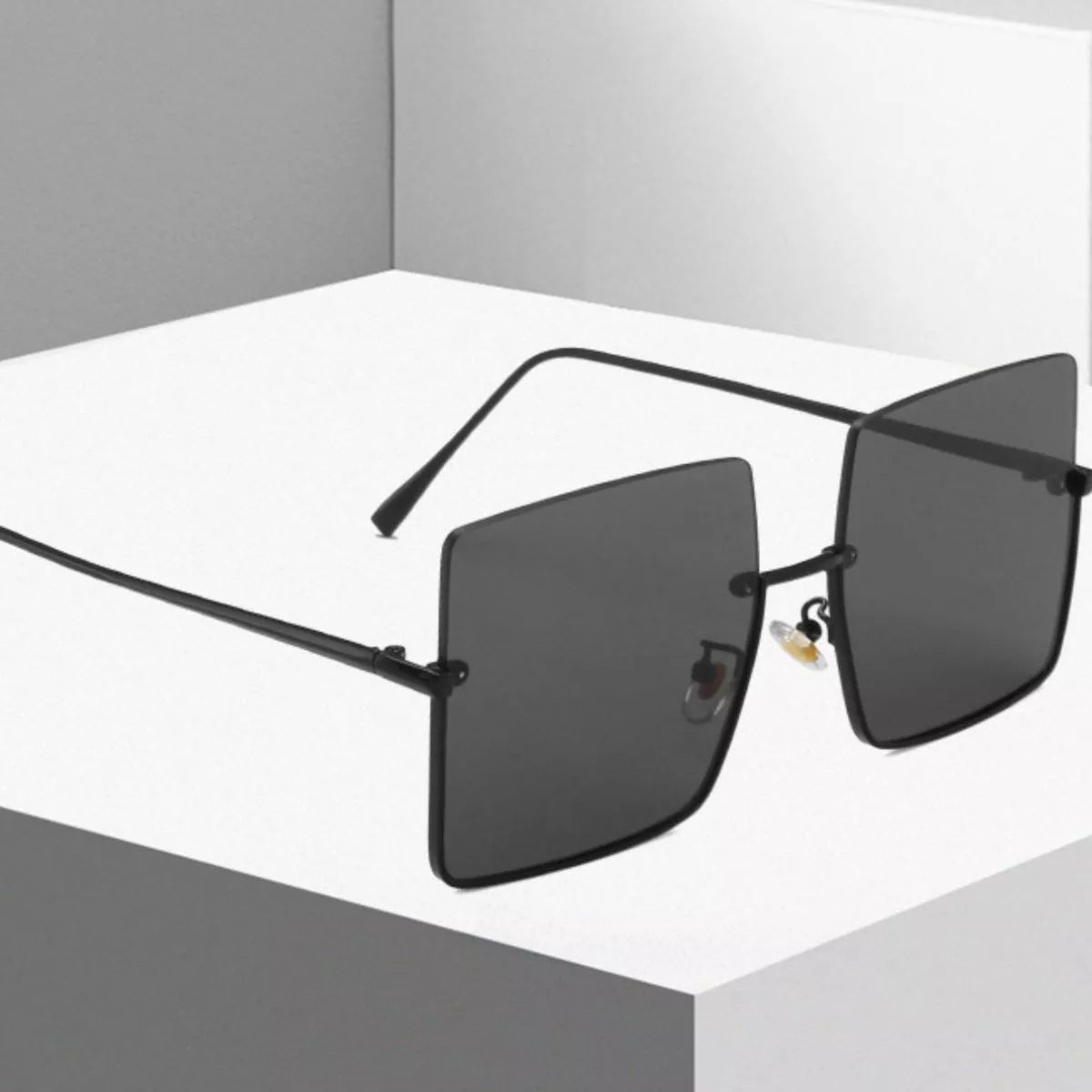 Black oversized square sunglasses with dark lenses, slim metal temples and visible nose pads displayed with open arms on a white pedestal