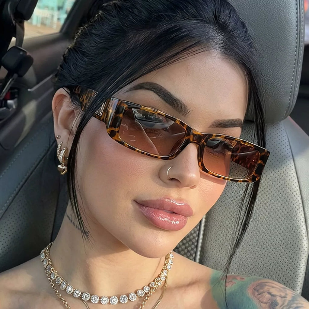 Woman wearing brown tortoiseshell-pattern rectangular sunglasses with brown gradient lenses, seated in a car with layered necklaces