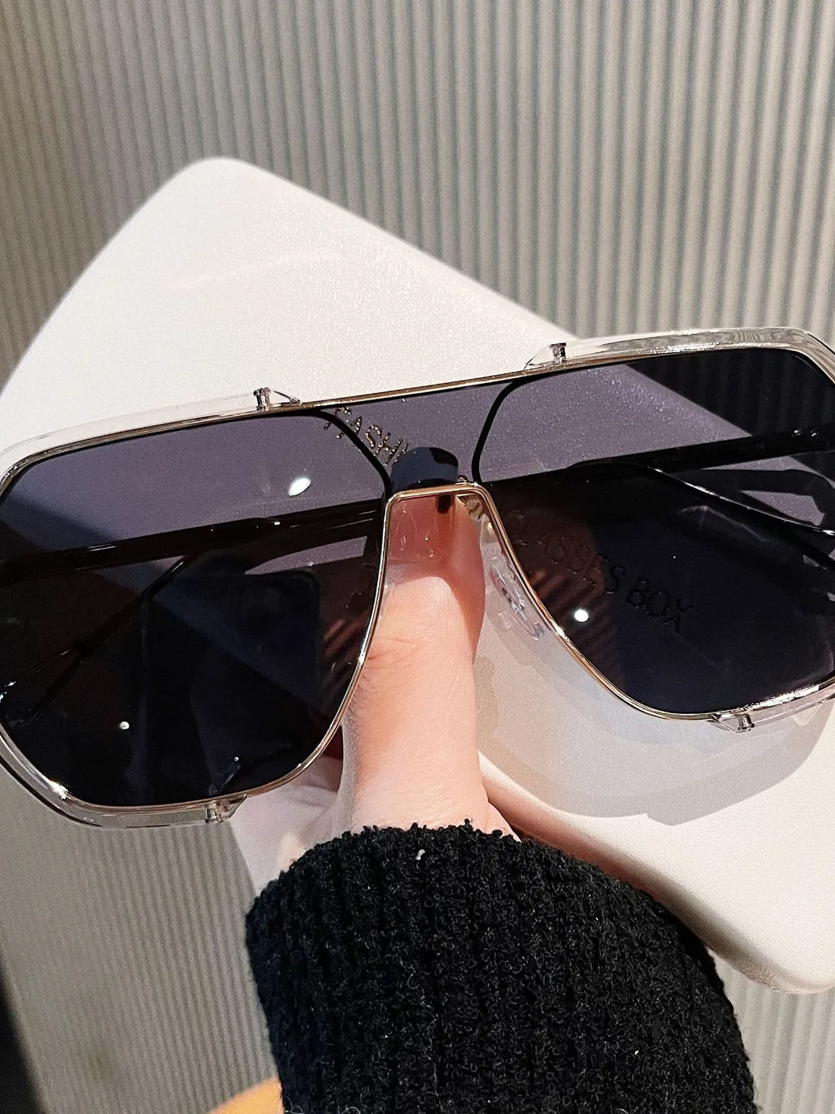Oversized aviator glasses with transparent-look rims, dark gray lenses, and a metallic double bridge, held over a white glasses box