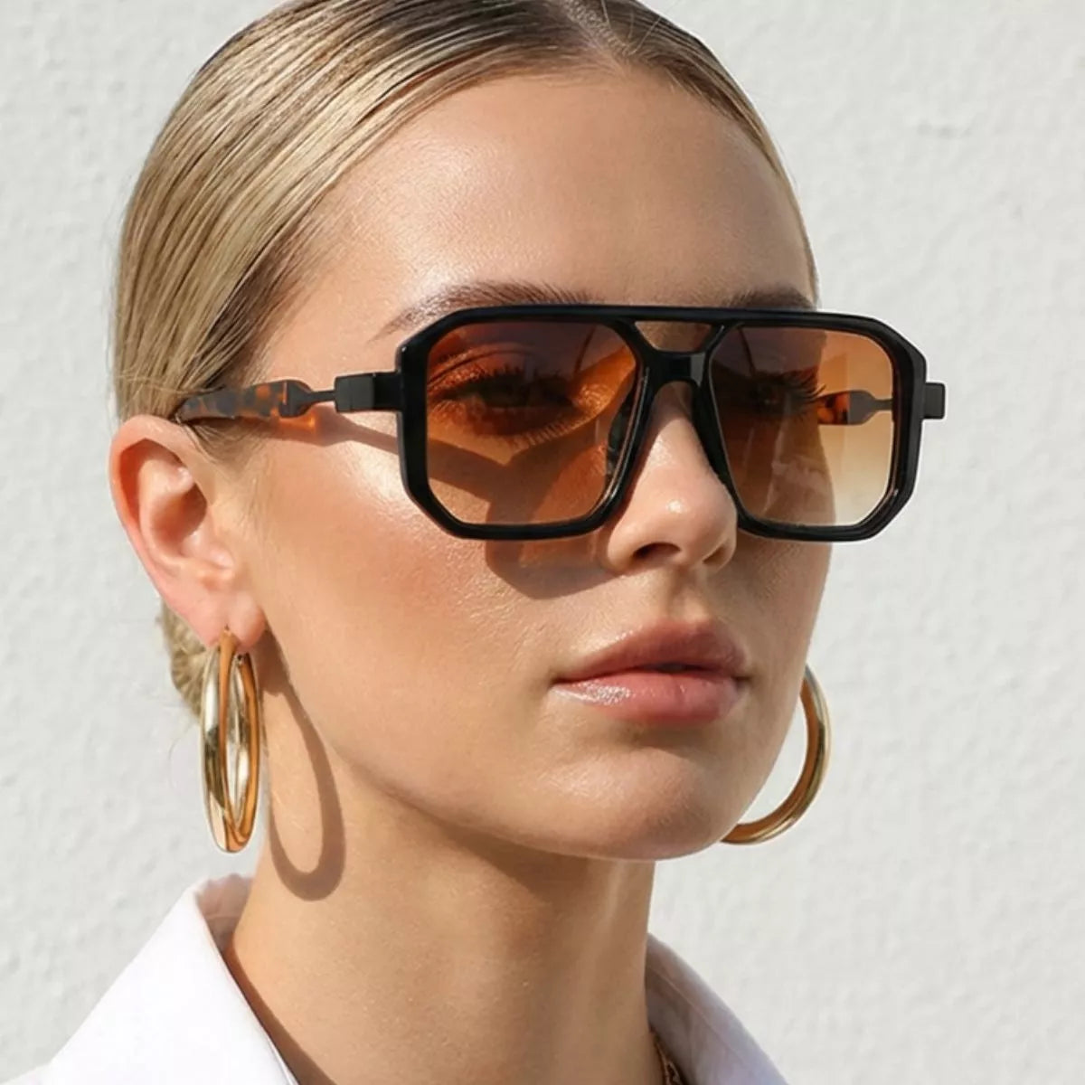 Model wearing black oversized square sunglasses with brown gradient lenses, gold hoop earrings and a white collared shirt against a light background