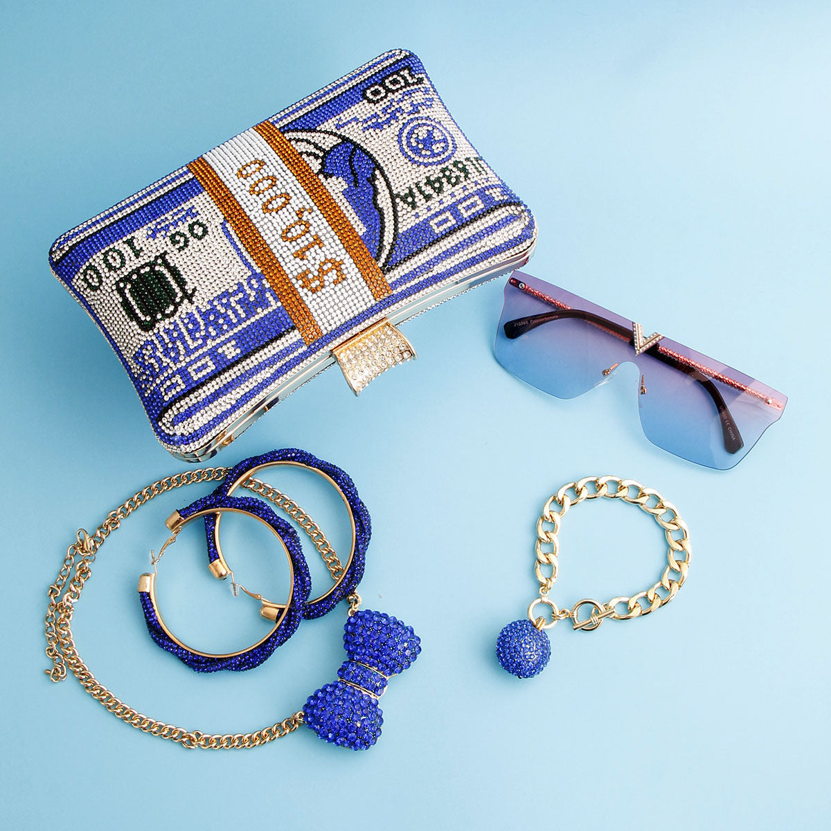 Blue rhinestone cash clutch bag with 100 dollar bill crystal design styled with blue rhinestone hoop earrings, gold chain necklace, gold curb chain bracelet with blue rhinestone ball charm, and blue tinted rimless sunglasses on light blue background