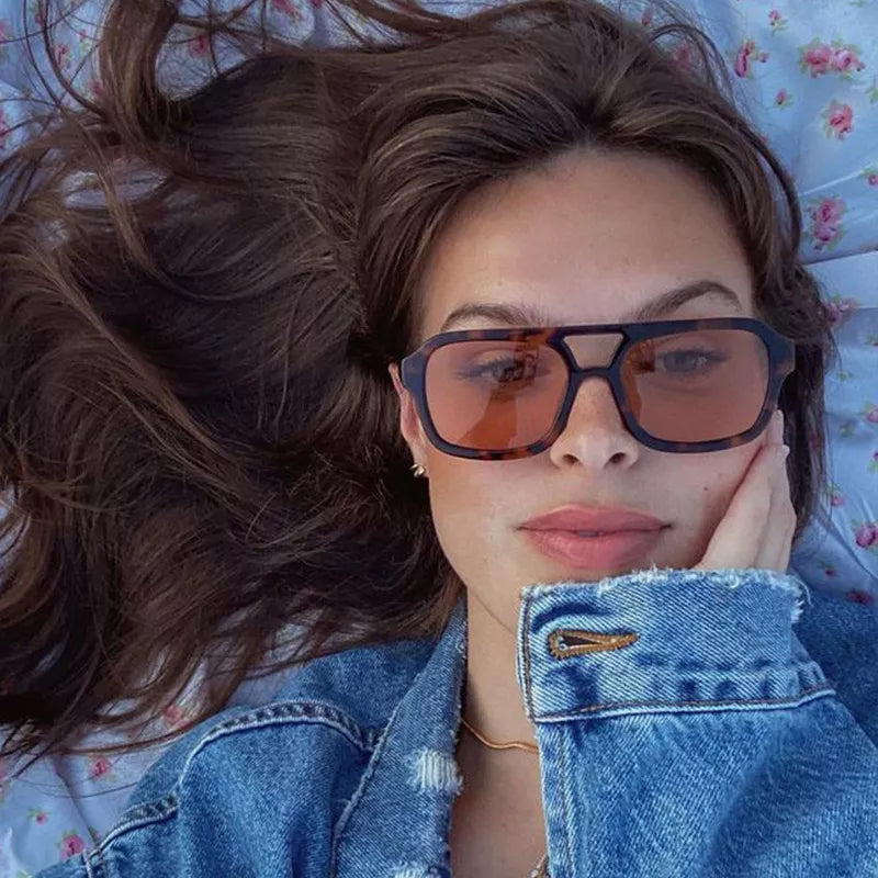 Woman wearing oversized tortoiseshell-pattern square sunglasses with warm-tinted lenses and a double bridge, lying on blue floral fabric in a denim jacket
