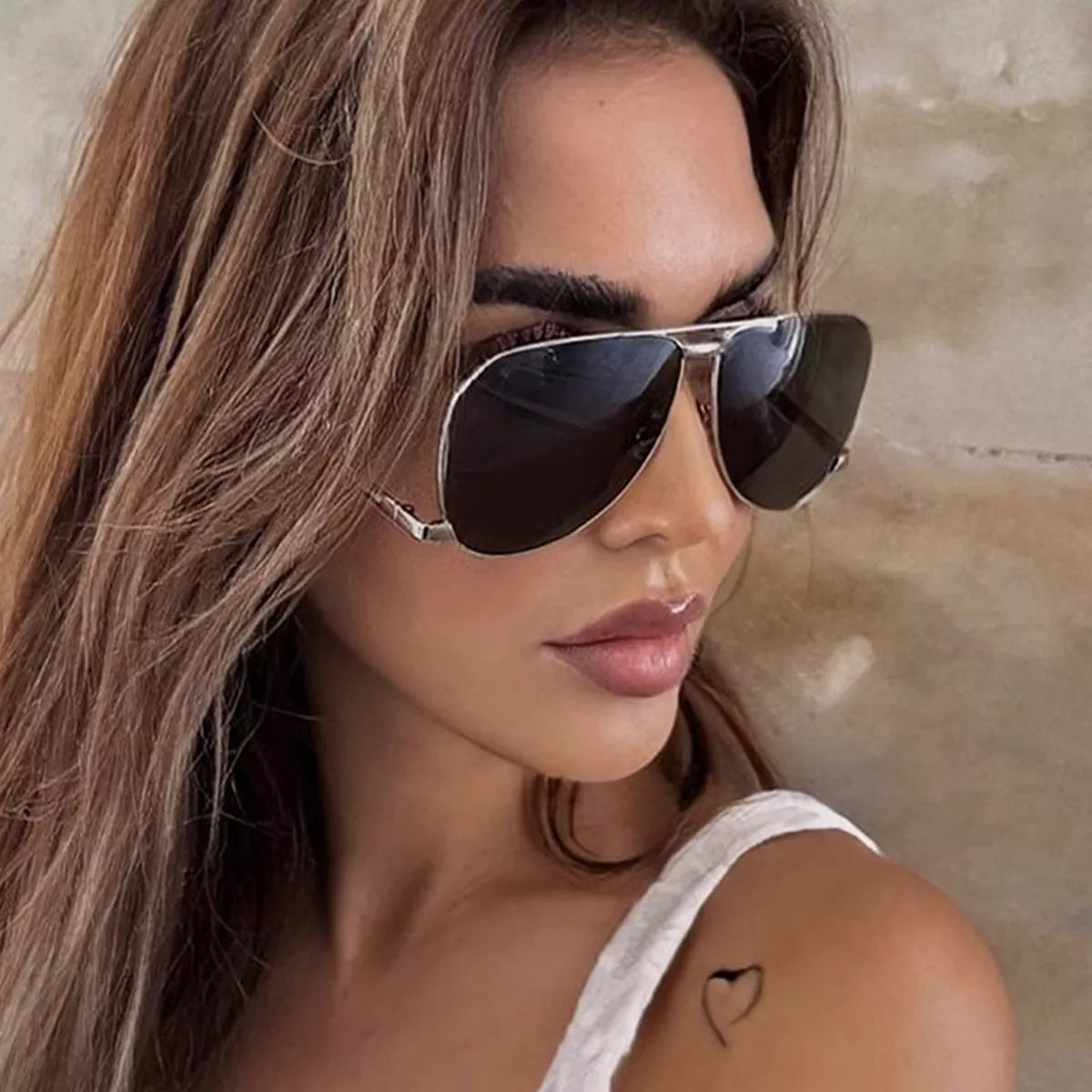 Woman with long brown hair wearing silver-tone aviator sunglasses with dark lenses and a white top against a beige stone wall