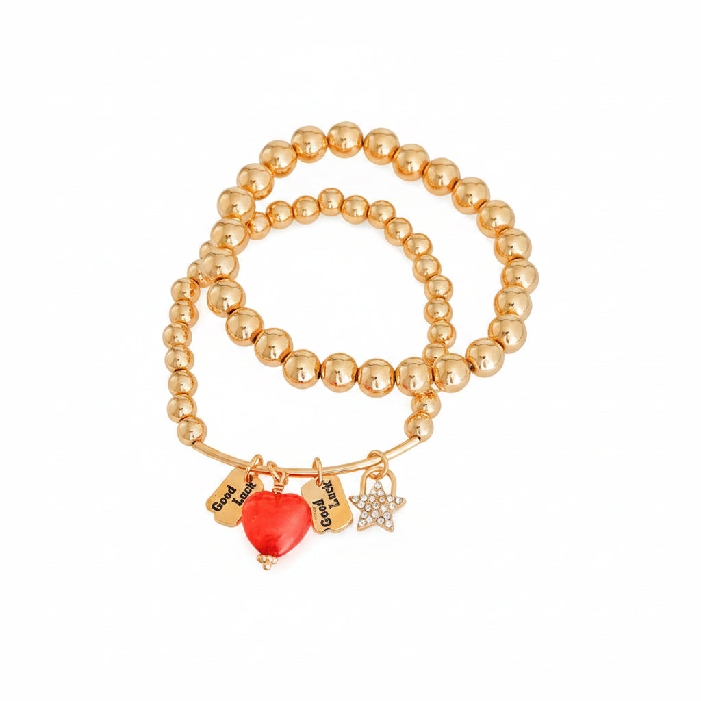 Two gold metal bead stretch bracelets stacked together — one with red enamel heart charm and two engraved Good Luck tags, one with rhinestone star charm and gold bar detail, white background