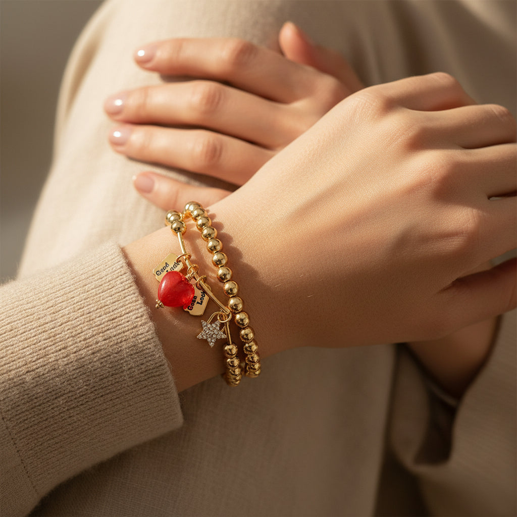 Two gold metal bead stretch bracelets stacked on woman's wrist over cream knit sleeve — red enamel heart charm, engraved Good Luck tags, and rhinestone star charm visible, warm neutral background