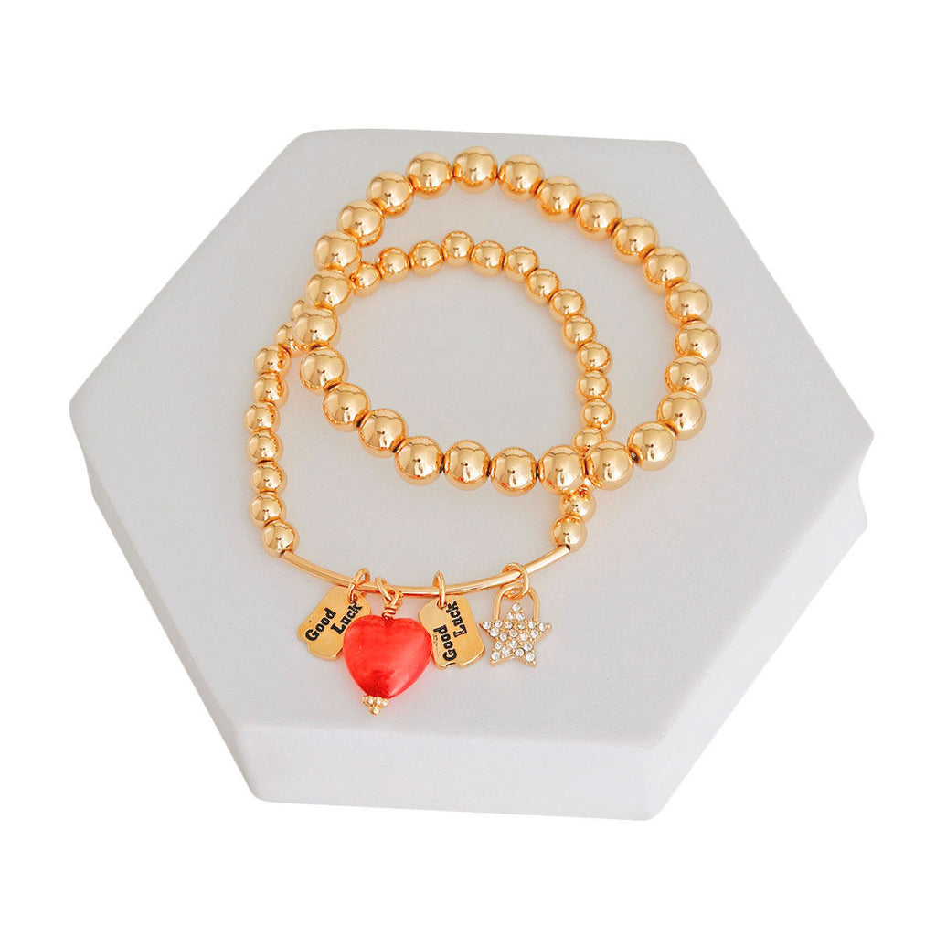 Two gold metal bead stretch bracelets coiled on white hexagonal jewelry display stand — red enamel heart charm with engraved Good Luck tags and rhinestone star charm with gold bar detail, white background