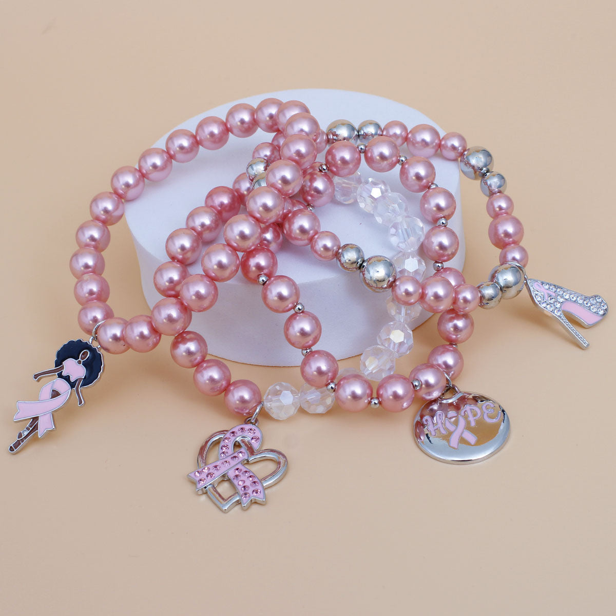 Four light pink pearl bracelets with iridescent clear and silver-tone beads draped over a white pedestal, showing pink ribbon Hope, heart, Afro woman and high heel charms