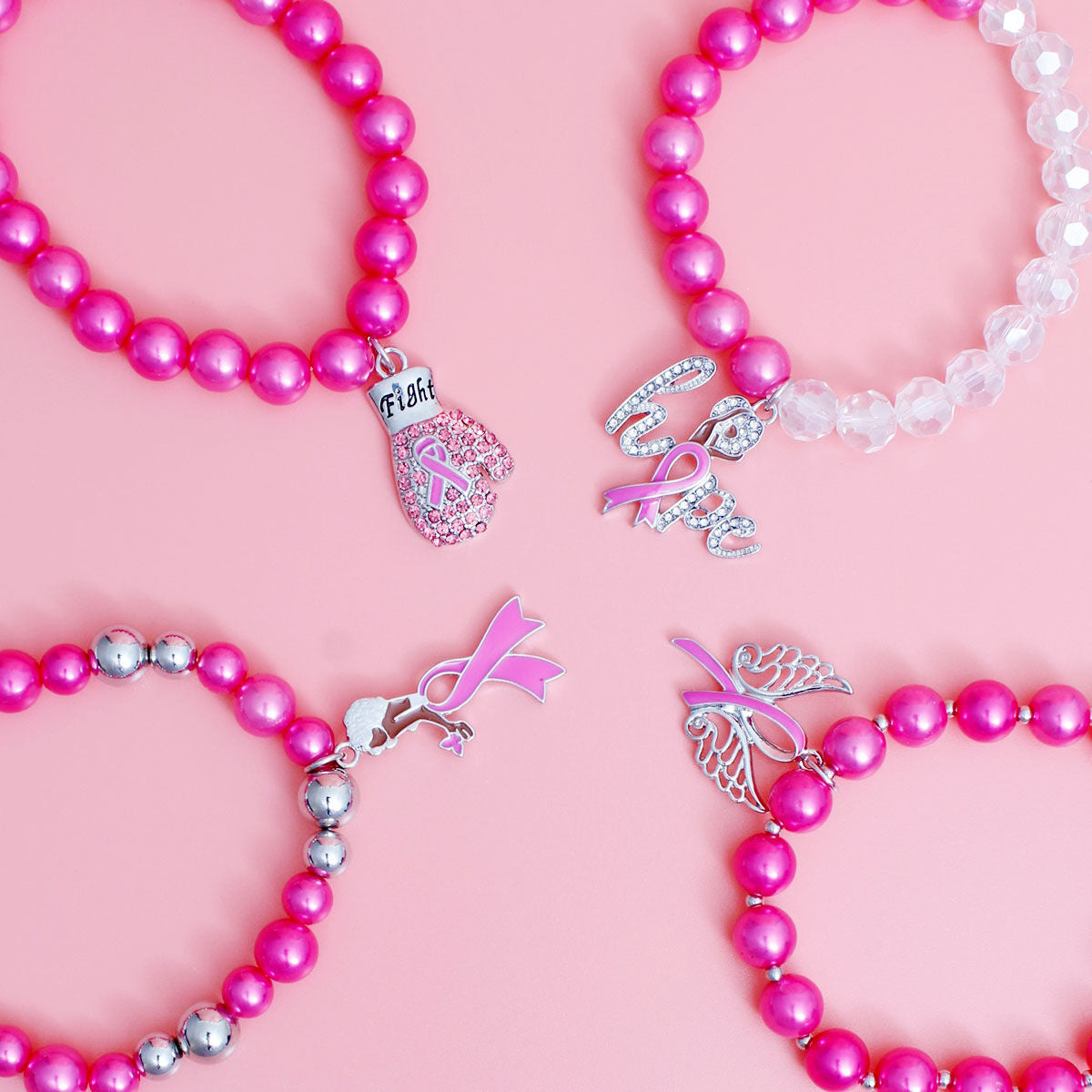 Four beaded breast cancer awareness bracelets arranged in a flat-lay on a solid pink background. The bracelets feature pink and silver beads, clear faceted beads, and silver charms shaped like a boxing glove with 'Fight' text, a ribbon with 'Love' text, a ribbon with a butterfly, and a ribbon with a heart.