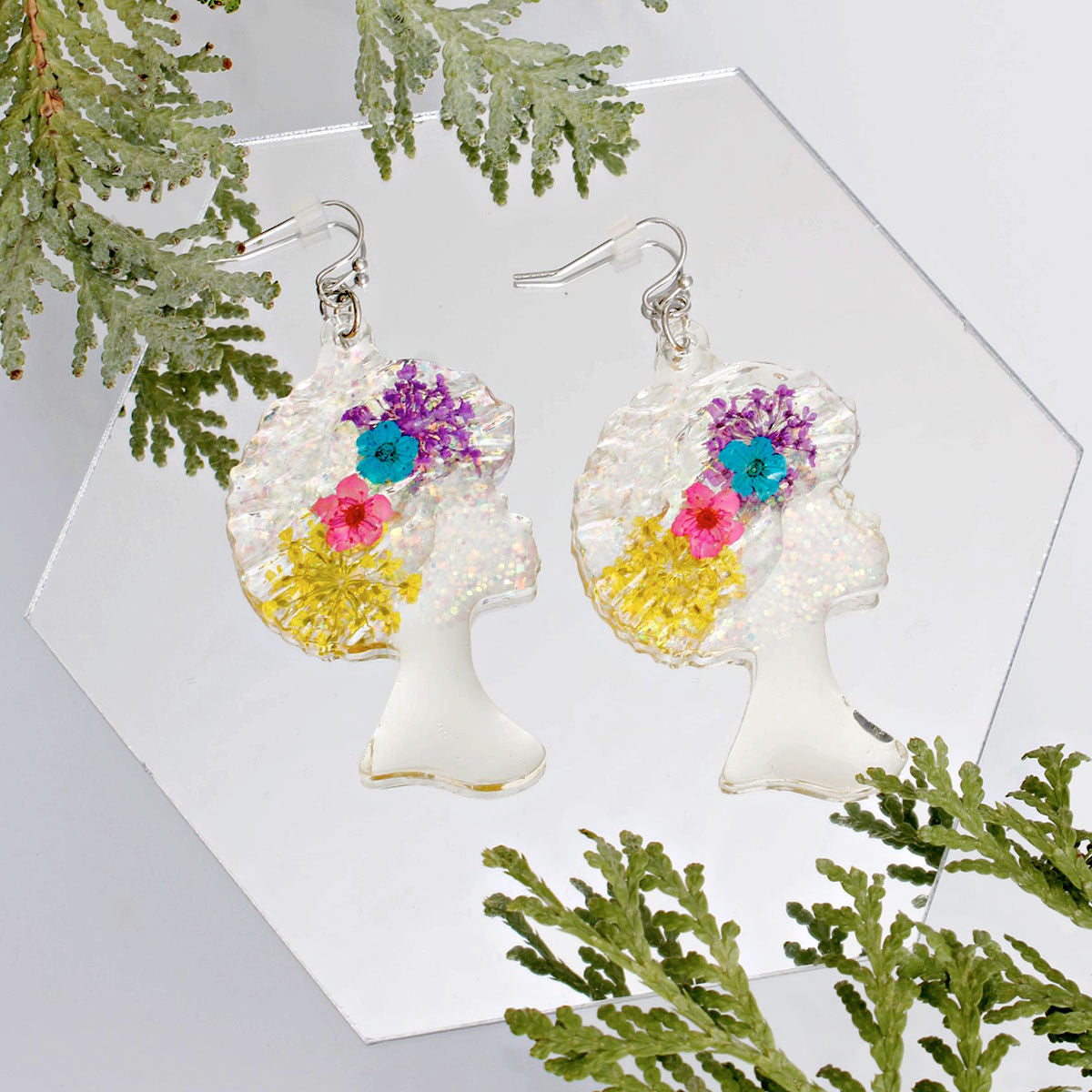 Pair of clear iridescent glitter acrylic Black girl afro silhouette dangle earrings displayed on white hexagonal mirror tile surrounded by green cedar branches, showing purple, teal, pink, and yellow multi-color flower details with silver fish hook wires