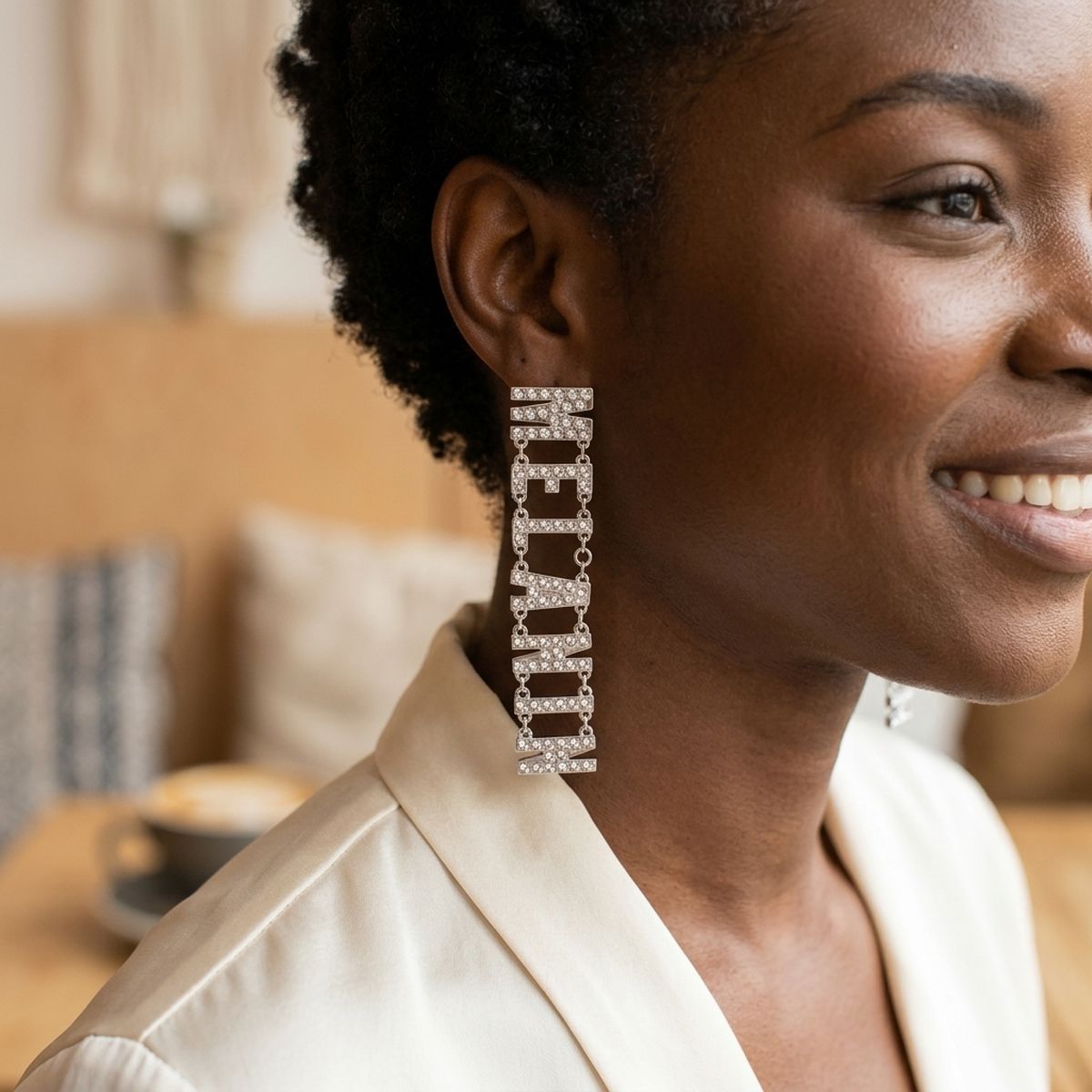 Smiling Black woman with short natural hair wearing silver clear rhinestone MELANIN linked letter dangle earring on post backing, shown in warm café setting wearing cream blazer
