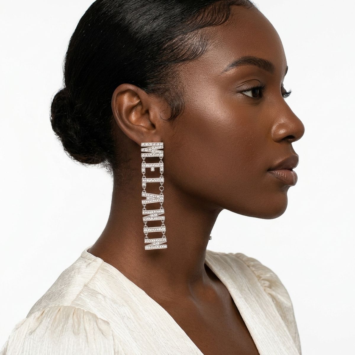 Side profile of Black woman with sleek low bun wearing silver clear rhinestone MELANIN linked letter dangle earring on post backing, shown against white background wearing white linen blouse