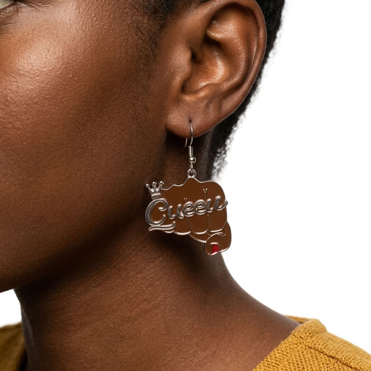 Close-up of Black woman wearing brown epoxy raised fist "Queen" crown dangle earring on silver fish hook wire, showing gold-tone script lettering and red nail detail against white background