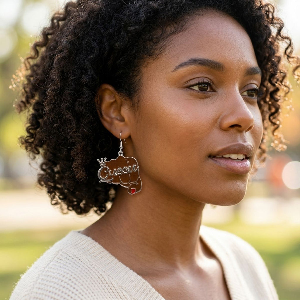 Black woman with natural curly hair wearing brown epoxy raised fist "Queen" crown dangle earring on silver fish hook wire, shown in outdoor natural light setting with blurred green background