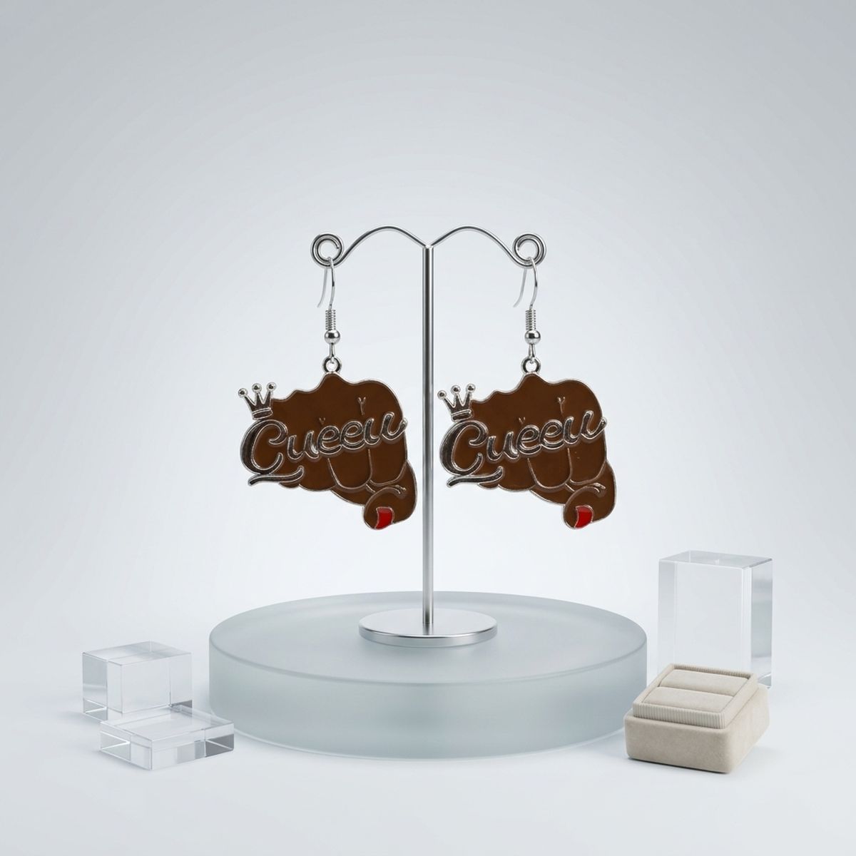 Pair of brown epoxy raised fist "Queen" crown fish hook dangle earrings hanging on silver double-hook earring display stand on frosted glass base, with clear acrylic blocks and cream velvet ring holder in background
