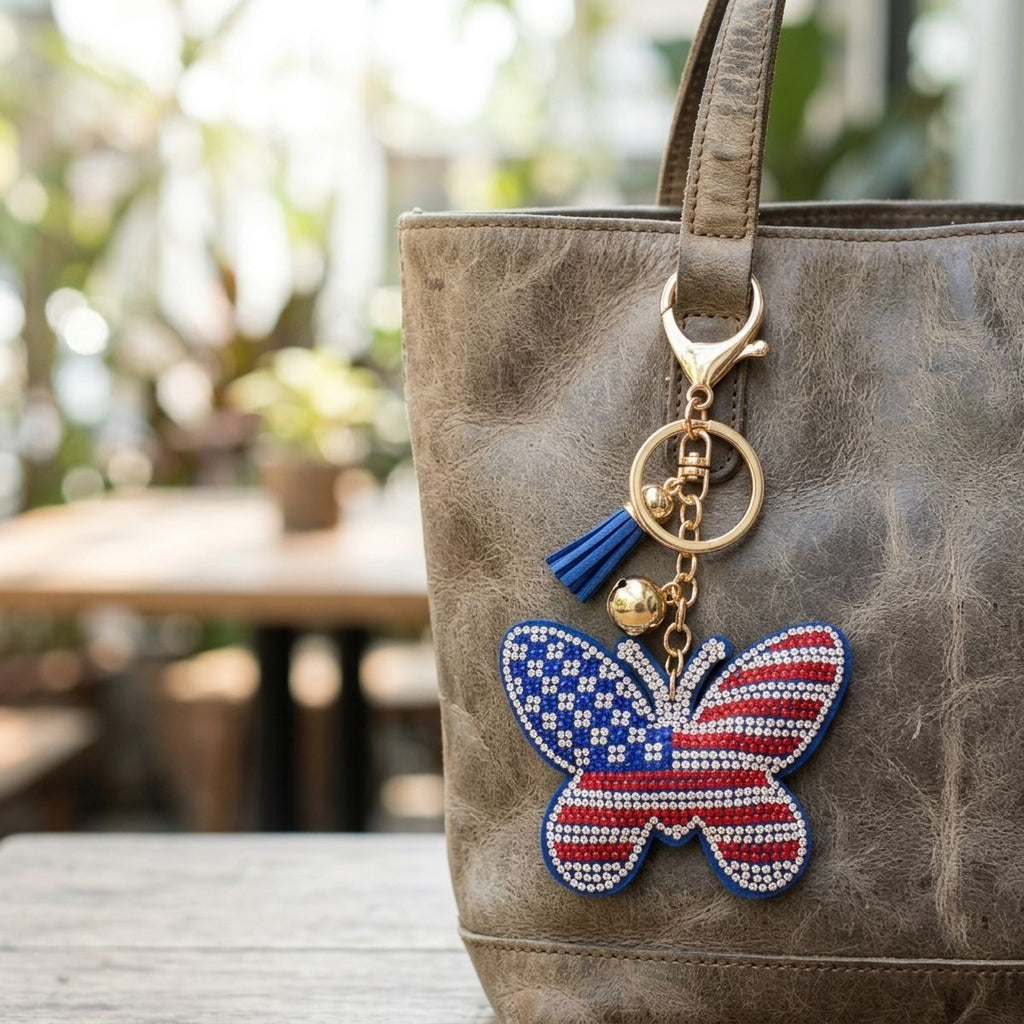 Patriotic butterfly rhinestone bag charm clipped to tan leather tote bag with blue tassel and gold hardware, outdoor setting