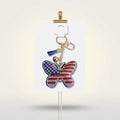 Patriotic butterfly bag clip keychain with American flag rhinestone detail, blue mini tassel, and gold lobster clasp on white display card