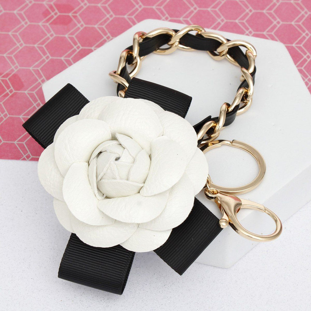 White 3D vegan leather camellia flower bag charm with black grosgrain ribbon bow and gold chain, shown attached to a car key fob with gold lobster clasp and split keyring on white background