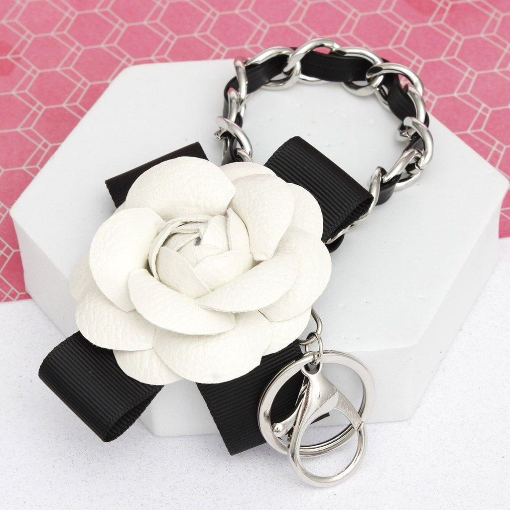 White 3D vegan leather camellia flower bag charm with black grosgrain ribbon bow and chunky silver chain woven with black faux leather, displayed on white hexagon stand with pink background