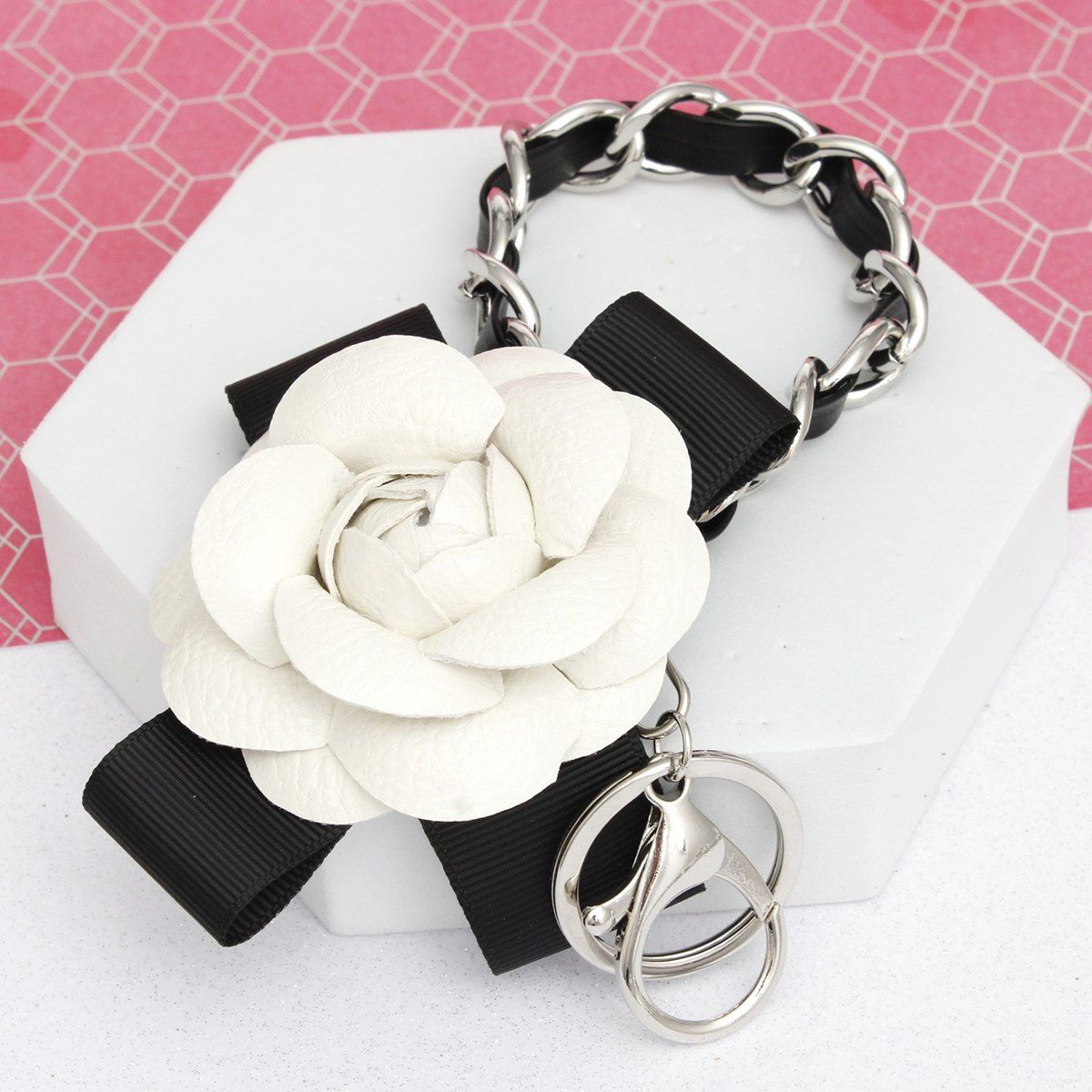 White 3D vegan leather camellia flower bag charm with black grosgrain ribbon bow and chunky silver chain woven with black faux leather, displayed on white hexagon stand with pink background