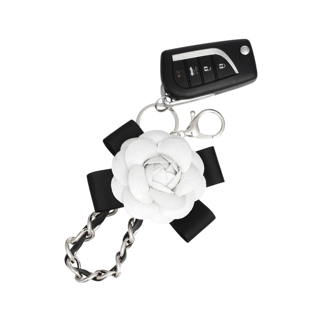 White 3D vegan leather camellia flower bag charm with black grosgrain ribbon bow and silver chain, shown attached to a car key fob with rhodium lobster clasp and split keyring on white background