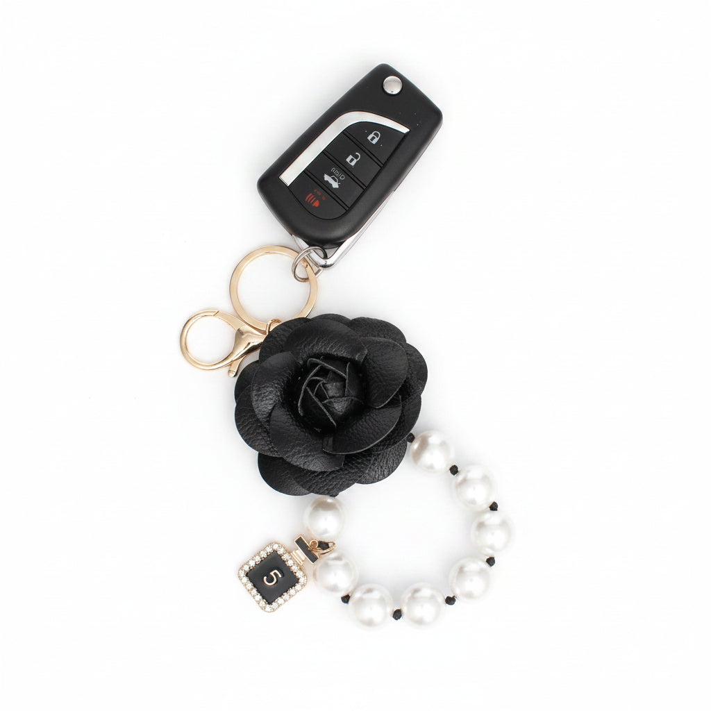 Black 3D vegan leather camellia flower bag charm with acrylic white pearl loop and mini gold perfume bottle charm, shown attached to a car key fob with gold lobster clasp on pink hexagon-patterned background