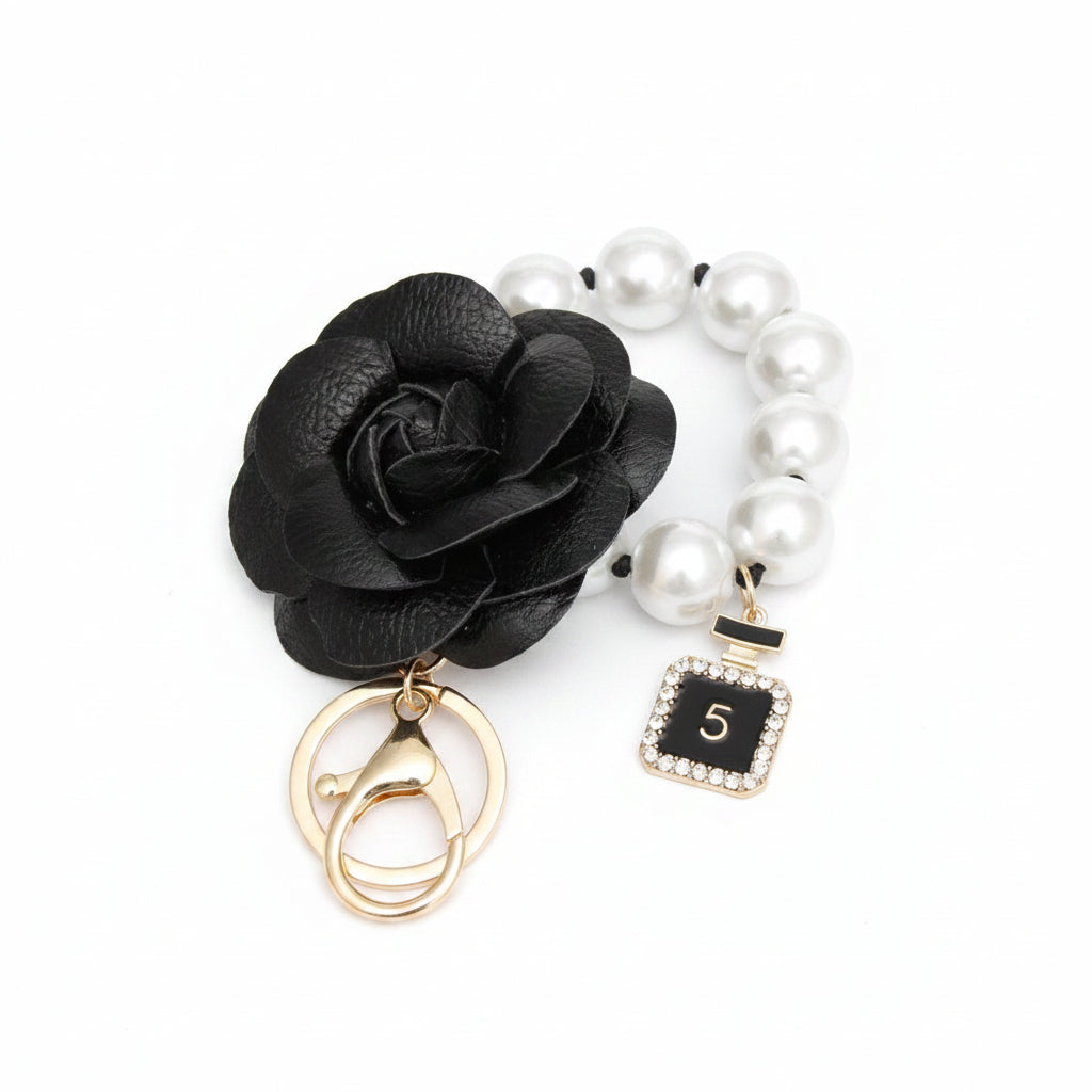 Black 3D vegan leather camellia flower bag charm with acrylic white pearl loop on knotted black cord and mini gold perfume bottle charm with black enamel and rhinestone border, displayed on white hexagon stand