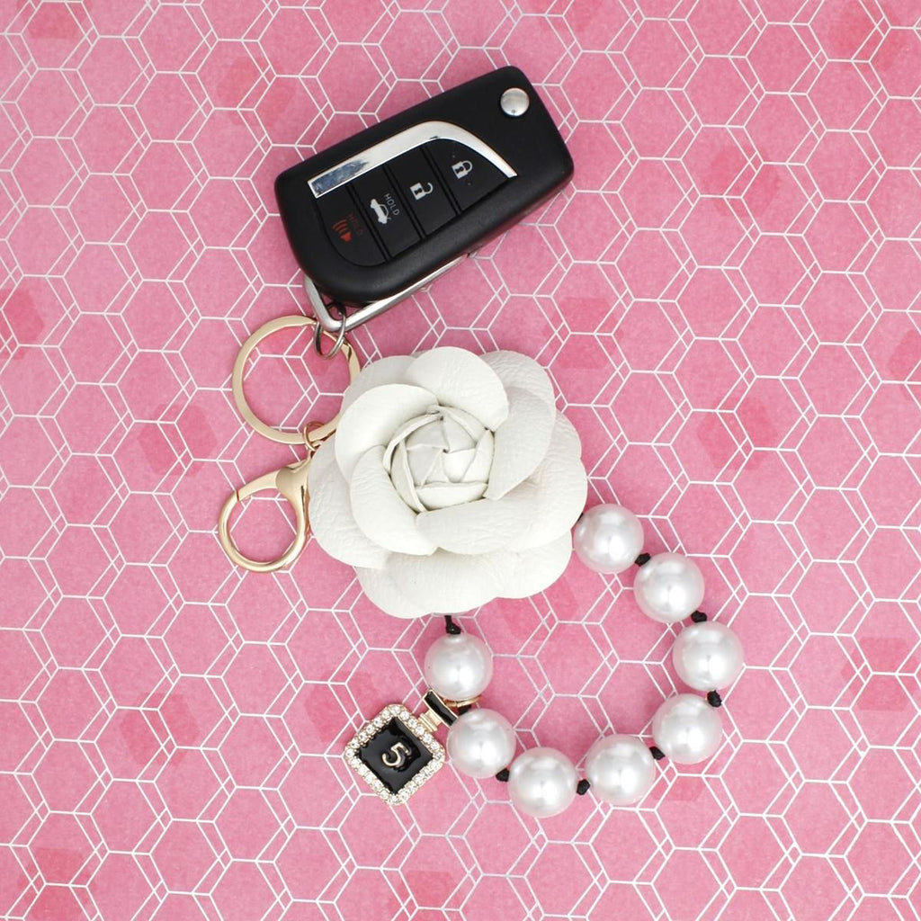 White 3D vegan leather camellia flower bag charm with acrylic pearl loop and mini gold perfume bottle charm with rhinestone detail, shown attached to a car key fob with gold lobster clasp and split keyring on pink background