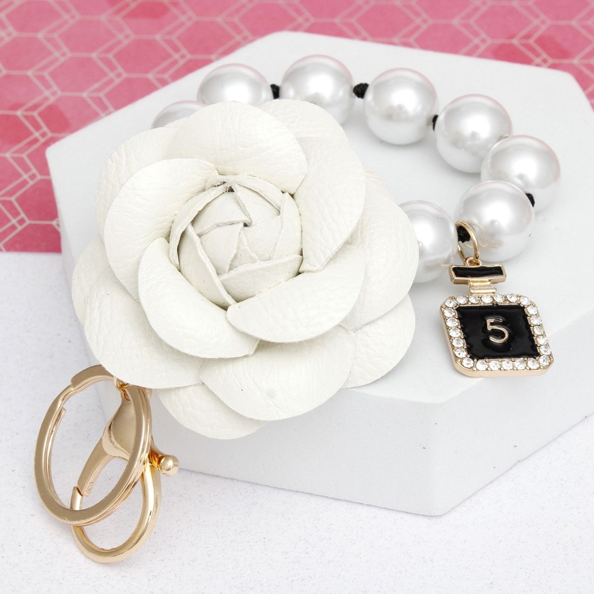 White 3D vegan leather camellia flower bag charm with acrylic pearl loop on knotted black cord and mini gold perfume bottle charm with black enamel and rhinestone border, displayed on white hexagon stand with pink background