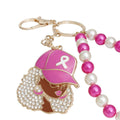 Close-up of breast cancer awareness keychain charm of a Black woman in a pink baseball cap with white ribbon, curly rhinestone hair, glasses, pink hoop earrings and pink lips, with gold lobster clip and fuchsia and cream pearl wristlet