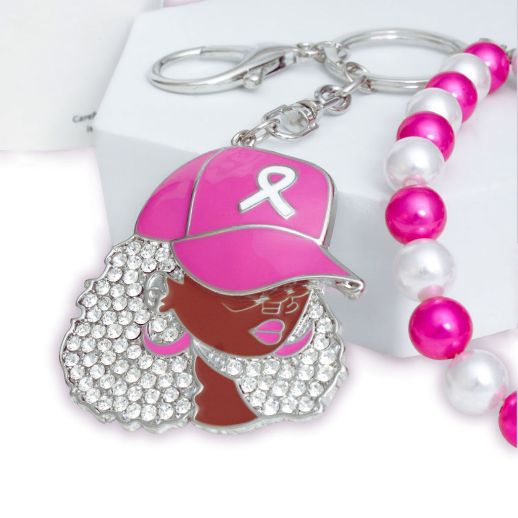 Close-up of silver-tone keychain charm of a Black woman in a pink baseball cap with white ribbon, curly rhinestone hair, glasses, pink hoop earrings and pink lips, beside fuchsia and white pearls