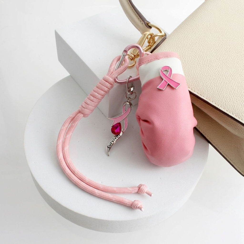 Pink ribbon boxing glove purse charm attached to a cream handbag with heart-shaped clip, survivor ribbon charm, crystal, and knotted cord