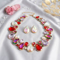 Light multicolor crystal collar necklace and matching teardrop stud earrings in gold-tone setting, displayed on white satin with pink rose and scattered crystals