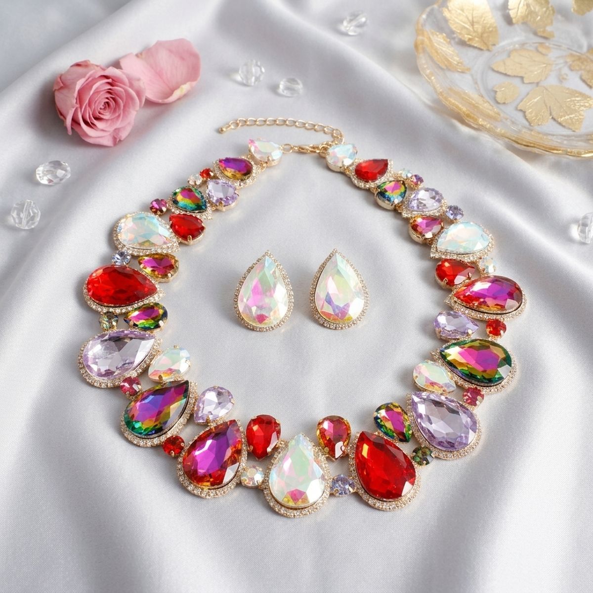 Light multicolor crystal collar necklace and matching teardrop stud earrings in gold-tone setting, displayed on white satin with pink rose and scattered crystals
