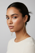 Female model with warm brown skin and dark hair in updo wearing large gold open diamond drop earrings with slim wire rhombus silhouette and gold fish hook wire, white ribbed sweater, gray background