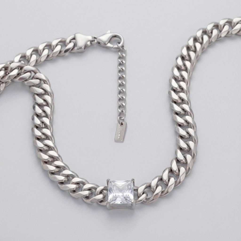Close-up overhead shot of an 8mm stainless steel Cuban curb chain necklace in silver arranged in a U-shape on a light grey surface. A single princess cut square cubic zirconia stone is bezel-set at the center bottom of the chain. A lobster clasp and 2-inch extender chain with a small rectangular stainless steel plate tag are visible at the top right.