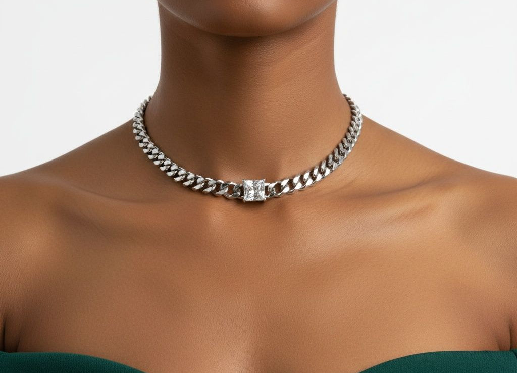 Close-up of a woman's neck and décolletage with dark skin wearing a deep emerald green off-shoulder dress against a white background. She wears an 8mm stainless steel Cuban curb chain necklace in silver sitting at the base of the neck, with a single princess cut square cubic zirconia stone bezel-set at the center front.