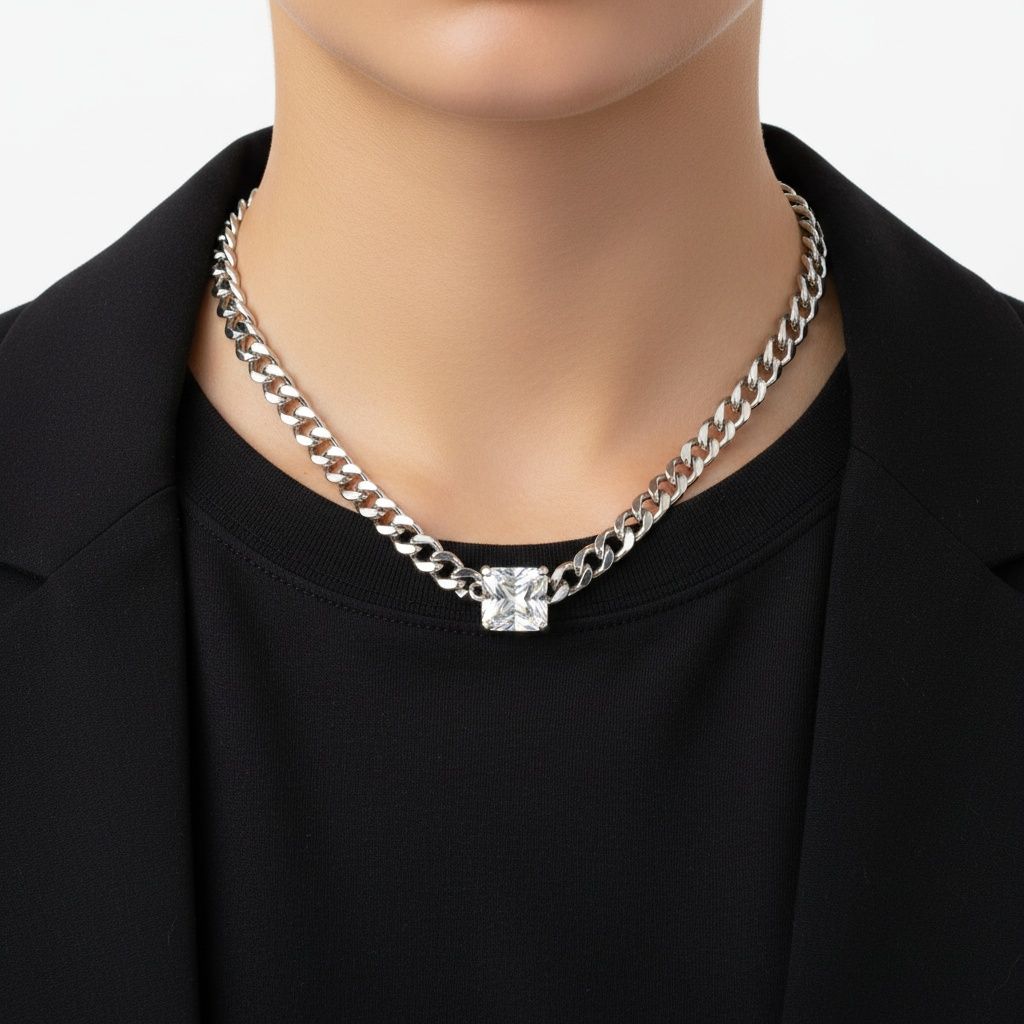 Close-up of a person's neck and chest wearing a black crew-neck top and open black blazer against a white background. They wear an 8mm stainless steel Cuban curb chain necklace in silver sitting at the collarbone, with a single princess cut square cubic zirconia stone bezel-set at the center front.